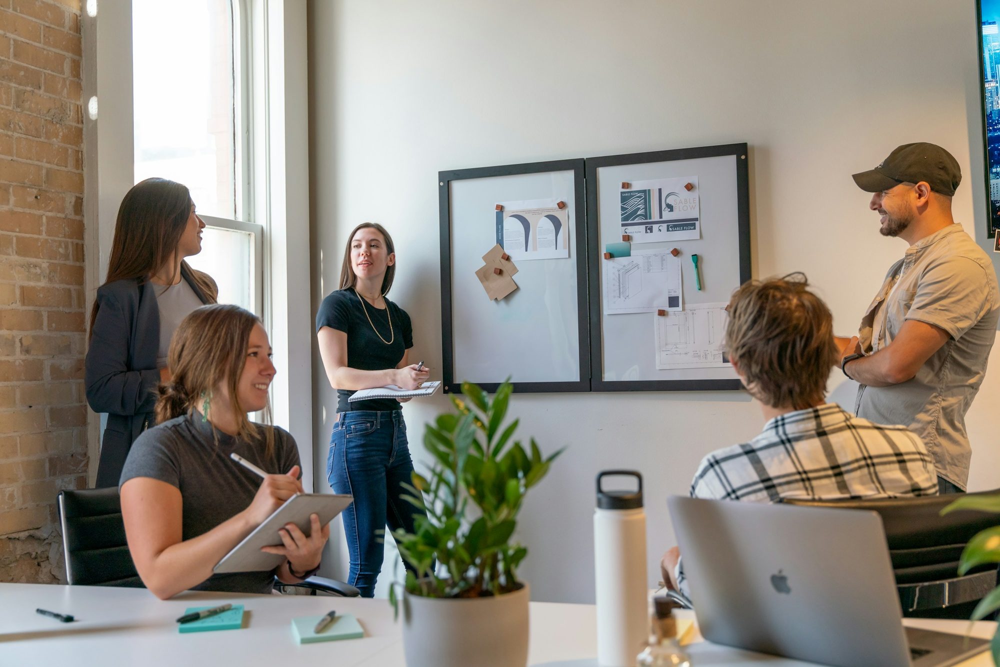 A product team meets in a sunlit office, one colleague taking notes on a tablet while others talk beside a pinboard of design sketches